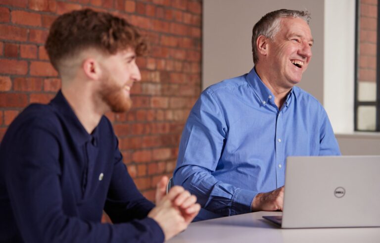 Man at desk laughing with another man smiling in foreground