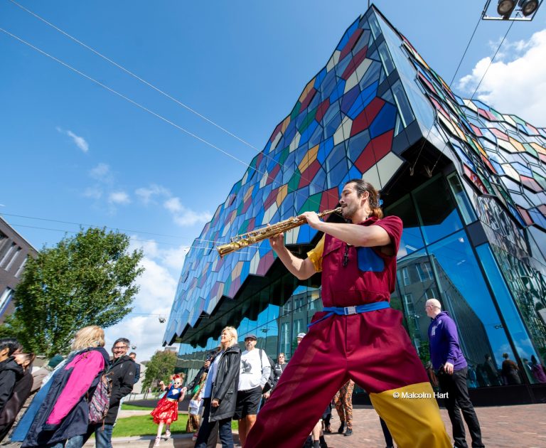 Man in red and gold clothing plays a trumpet in front of a colourful building.