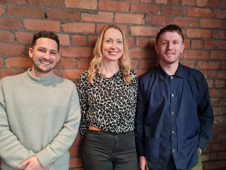 Two men and a woman smile standing in front of a brick wall
